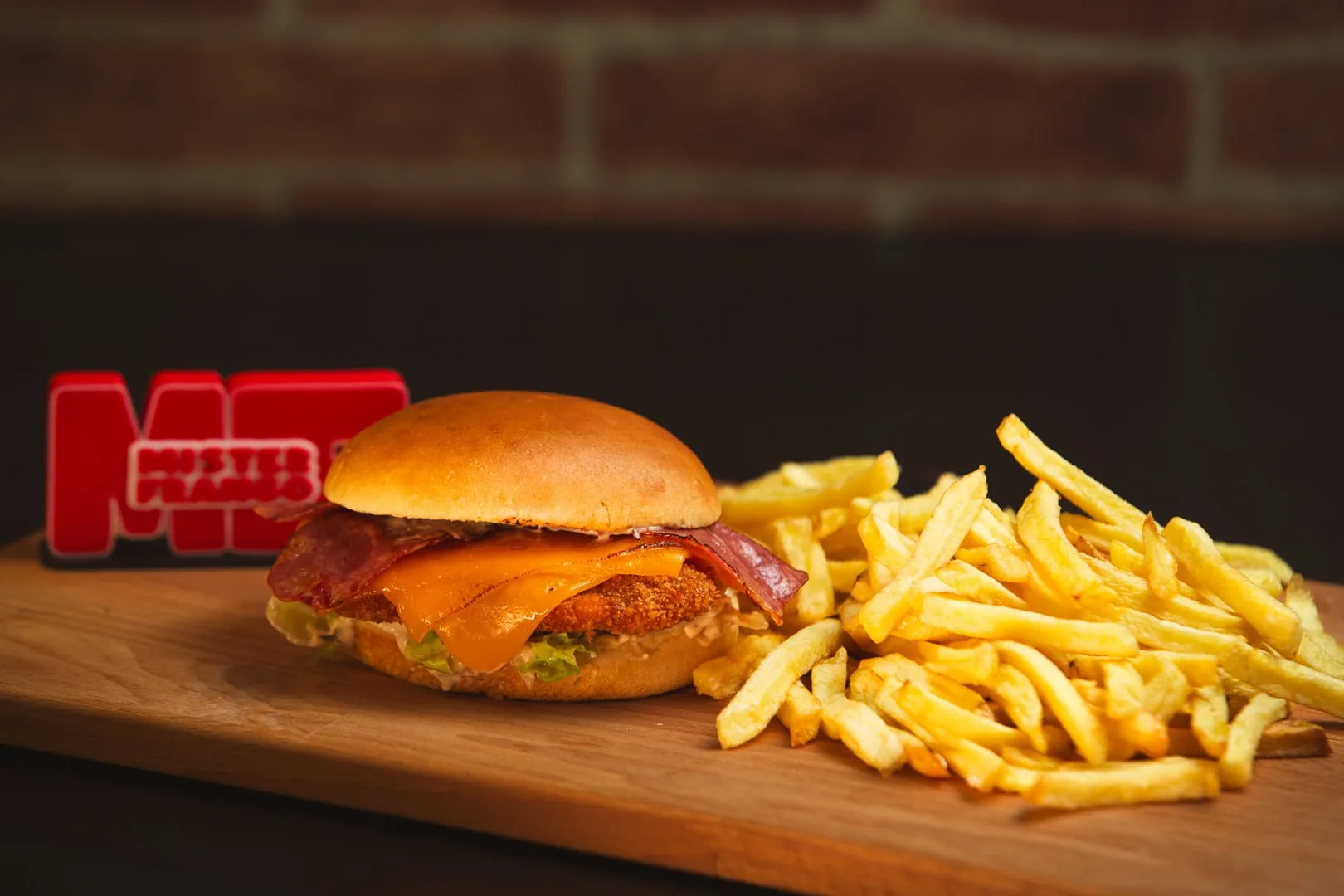 Sanduíche Mister Crispy com bacon e queijo, ao lado de batata frita numa tábua de madeira, com o letreiro néon do Mister Frango ao fundo